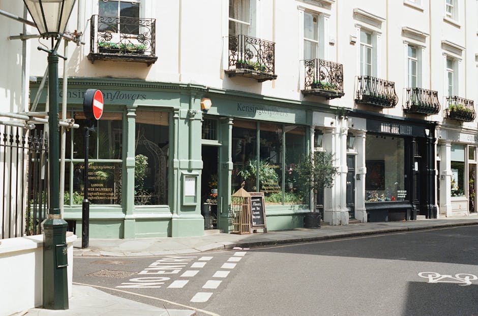 A florist shop with a light green exterior featuring large glass windows displaying floral arrangements inside. The storefront has a black sign and a small chalkboard sign on the sidewalk advertising 