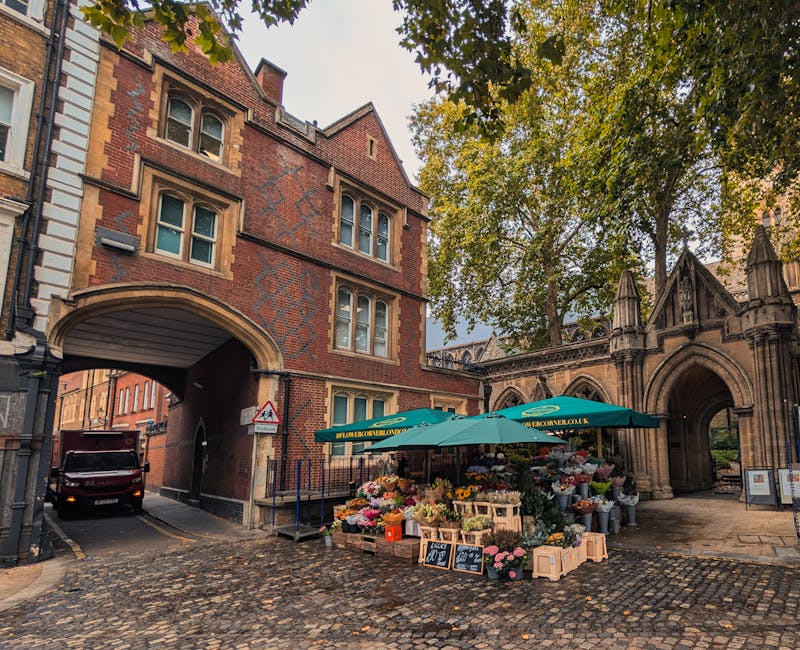 A flower stall set up on a cobbled street beneath an archway of a historic red-brick building, featuring a large green umbrella with the floriststjohnswood.co.uk logo. The stall displays an abundant s
