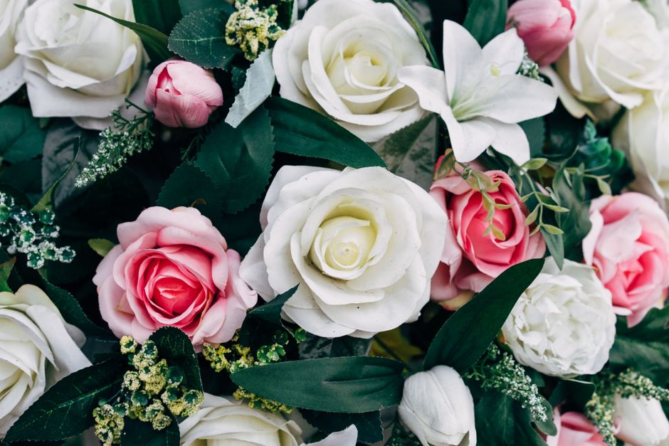 A close-up view of a floral arrangement featuring a bouquet of fresh roses in shades of white, blush pink, and deeper pink, complemented by white lilies and delicate green foliage. The bouquet is arra