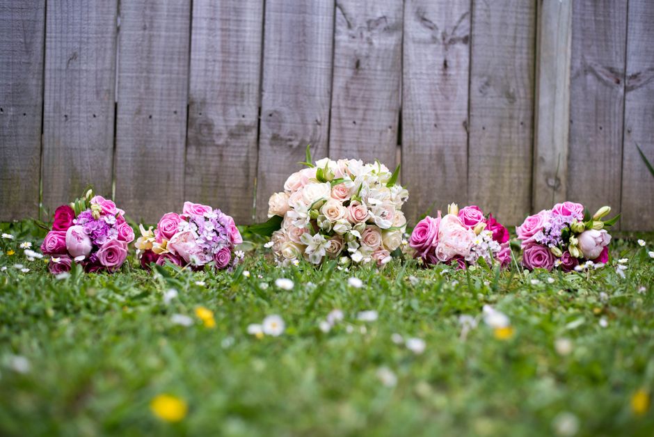 A collection of five floral bouquets arranged on a grassy surface outdoors in front of a wooden fence. The bouquets include pink, purple, and cream-colored roses, with some white lilies and hydrangeas