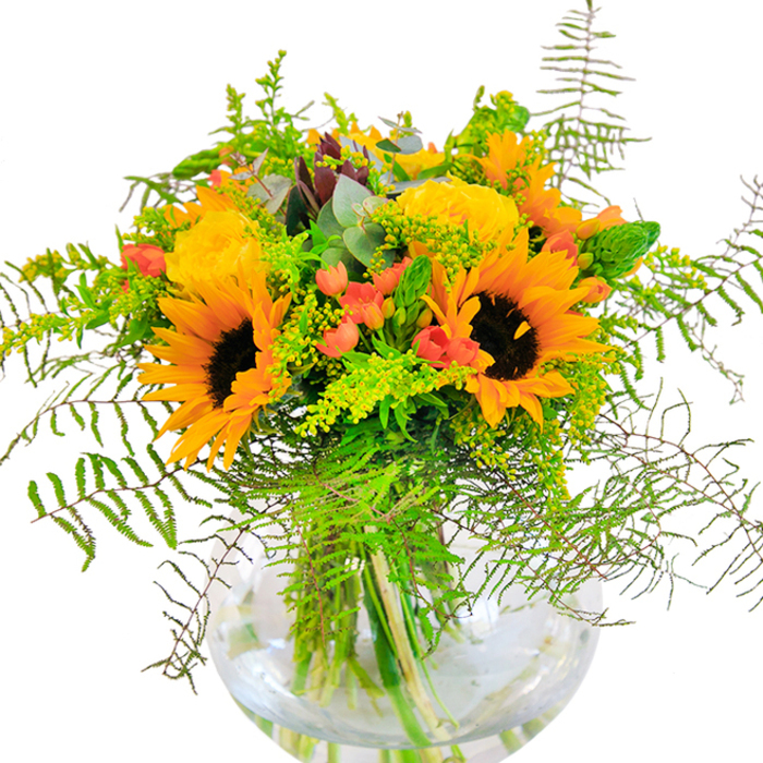 The first impression of this stunning bouquet is one of sunlit celebration-a vibrant ode to the bright, cheerful days in St John's Wood. Sunflowers take centre stage, their golden petals unfurling in arcs around velvety, dark centres that anchor the arrangement with warmth and strength. Filling the spaces between, tightly packed yellow garden roses open slowly, their spiral forms recalling the elegant blooms found in the area's private gardens. Splashes of rich red Hypericum berries add instant energy, their glossy skins a joyful surprise among the soft flower heads and trailing greenery. Fern fronds and wispy goldenrod breathe movement into the composition, spilling outward like verdant waves inspired by the gentle landscape along Hamilton Terrace. Slender green spikes rise among the foliage, and the occasional eucalyptus leaf-muted and blue-green-peeks through, granting a rustic authenticity. All these elements gather in a sparkling, spherical glass vase, its transparency revealing fresh, robust stems arranged in cool water. The piece is alive with colour, subtle scent, and texture-a bouquet destined for marking treasured occasions, gifting delight, or uplifting a room just steps from St John's Wood Church Gardens. The artistry is unmistakable, reflecting both the florists' deep local roots and the sophisticated character of NW8.