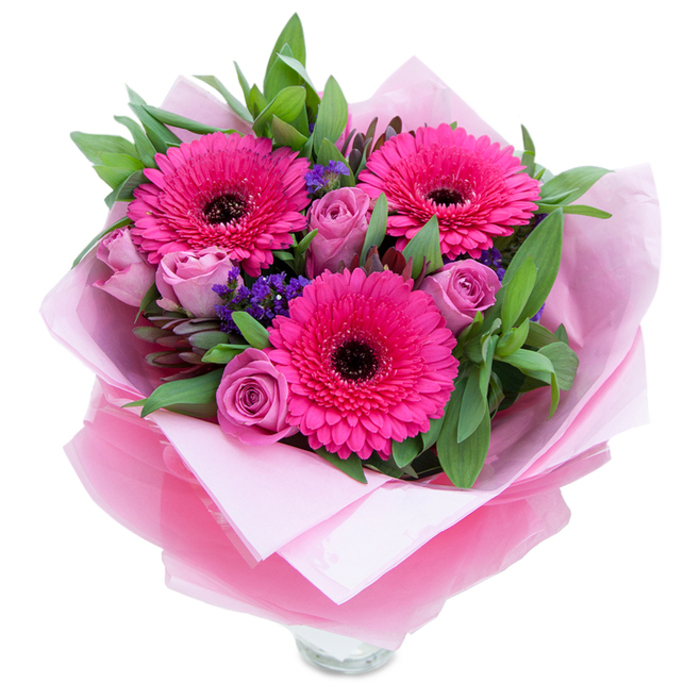 I picture this bundle of blooms as a tender moment shaped into a bouquet, the sort I'd prepare for a customer collecting flowers after a stroll past Abbey Road or the peaceful walks near Wellington Place. In the centre, three fuchsia-pink Gerbera daisies open boldly, each petal layered in concentric rows that catch the light and direct the eye inward to a dark, almost black core. Around them, roses in a dusty lavender-pink remain gently furled, their compact cups promising silk-soft layers as they open; these blooms add a subtle, romantic softness to the arrangement. Tiny sprays of purple statice slip between the larger flowers like little islands of colour, their texture crisp and slightly papery against the velvety roses. The greenery-broad salal-style leaves or hints of eucalyptus-sits full and fragrant behind the blooms, contributing a fresh, clean aroma and a structural rhythm to the silhouette. The bouquet is presented with care: a base of pale pink matte tissue cradles the stems, topped by a layer of translucent fuchsia-pink cellophane that shimmers and folds with a handcrafted finish; the glass vase rim peeks out at the bottom, ready for water and immediate enjoyment. The overall impression is one of intimate celebration: a thoughtful arrangement suited to birthdays, anniversaries, or a quiet 'thank you' in the St John's Wood neighbourhood, crafted to convey warmth, attention to detail, and floral longevity.